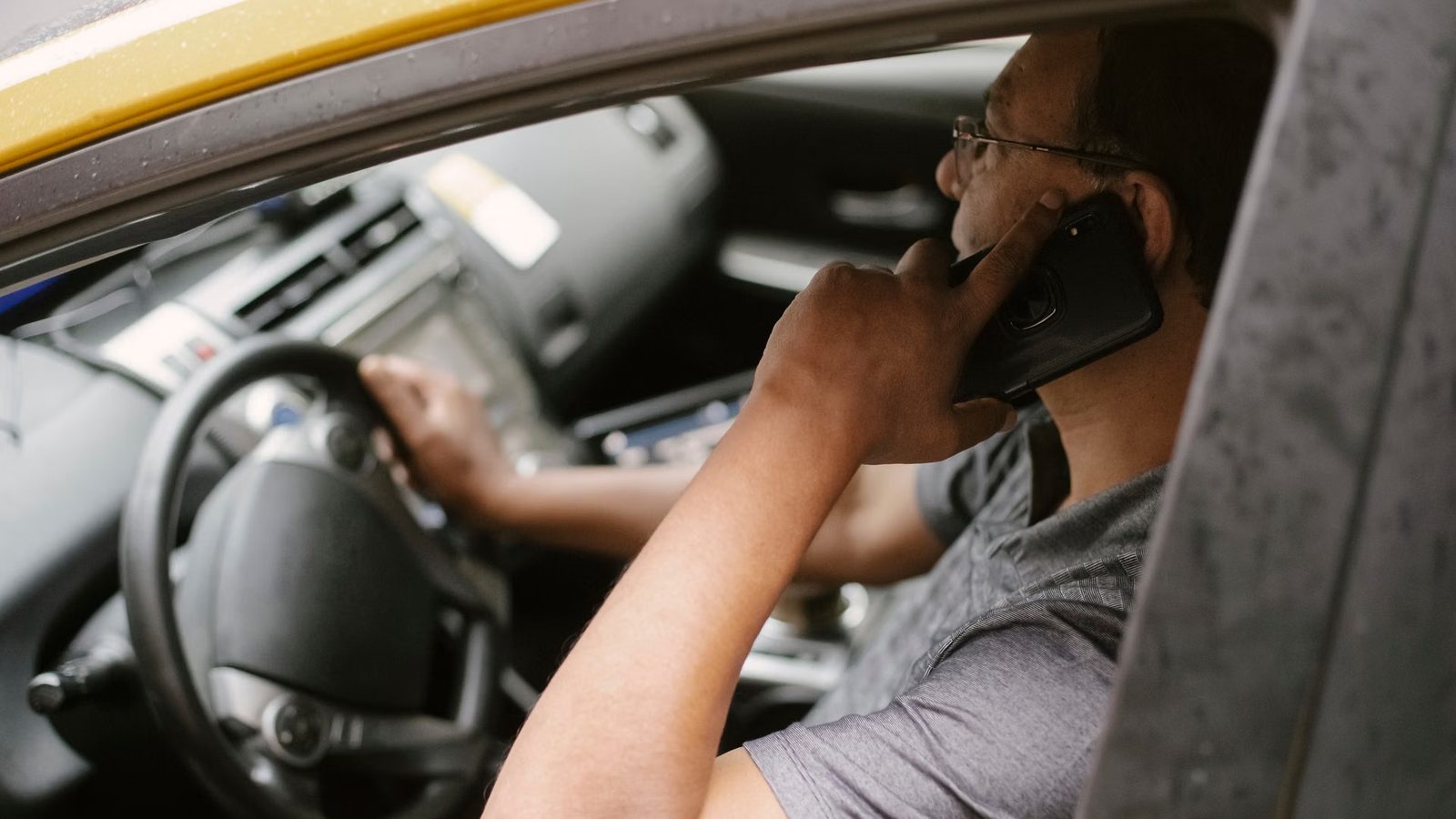 Mężczyzna w okularach rozmawia przez telefon komórkowy, siedząc za kierownicą samochodu z opuszczoną szybą.