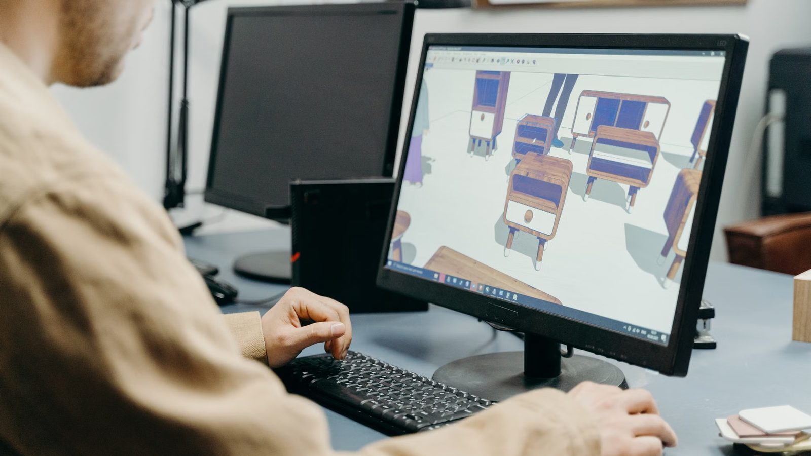 Mężczyzna przy biurku przegląda na monitorze trójwymiarowy projekt drewnianych szafek, obok drugi monitor i klawiatura