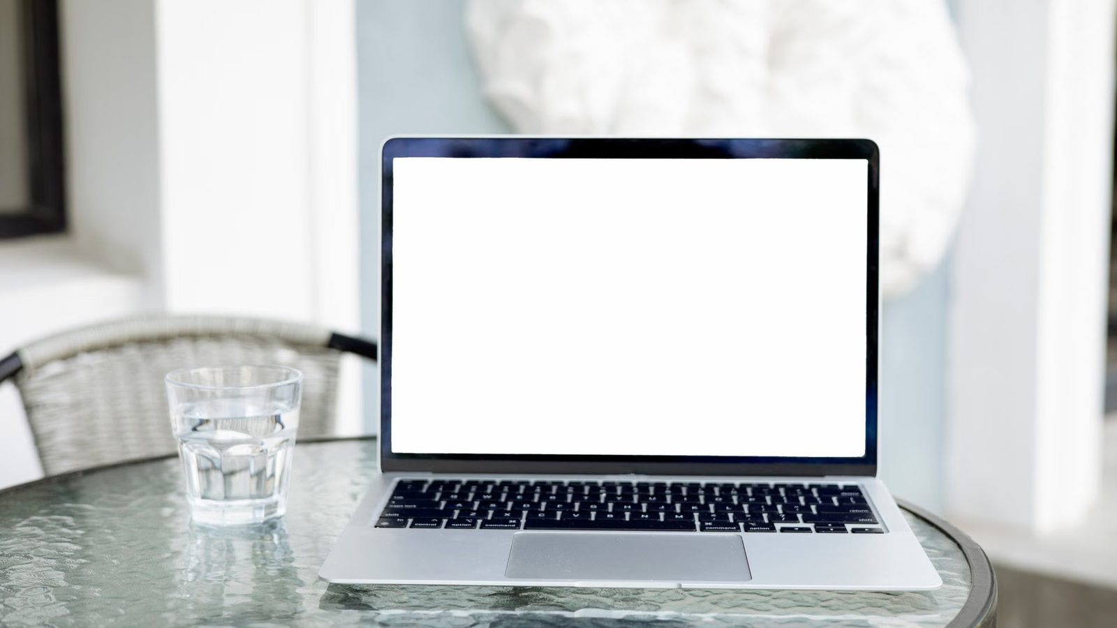 Otwarty laptop z pustym jasnym ekranem stoi na szklanym stoliku obok szklanki z wodą