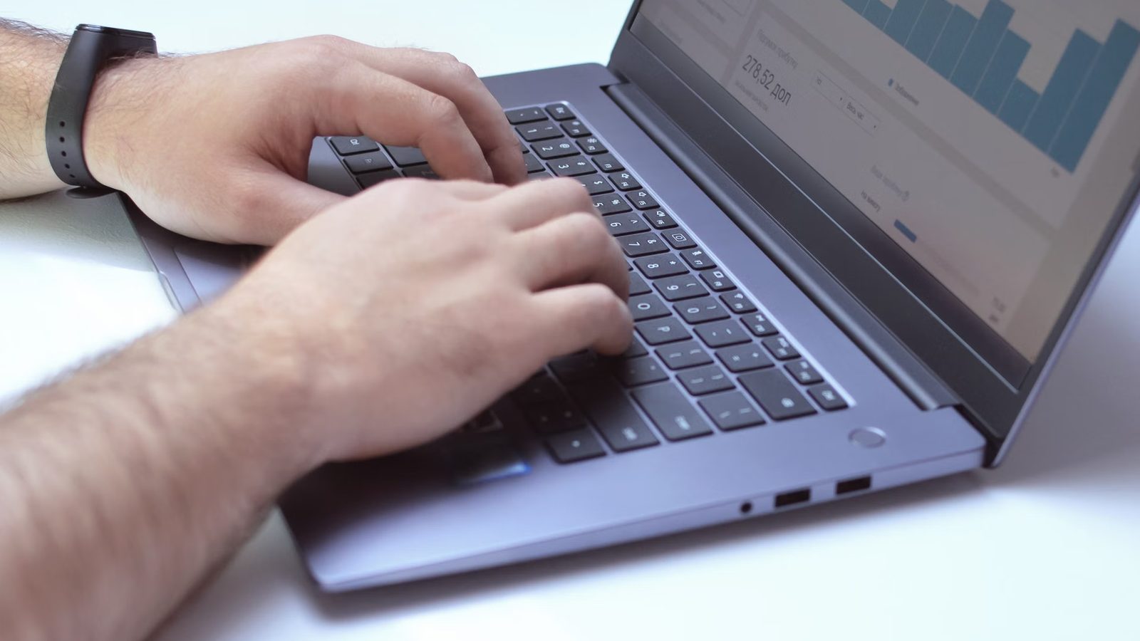 Dłonie mężczyzny piszącego na klawiaturze laptopa, na ekranie wykres słupkowy i panel z danymi