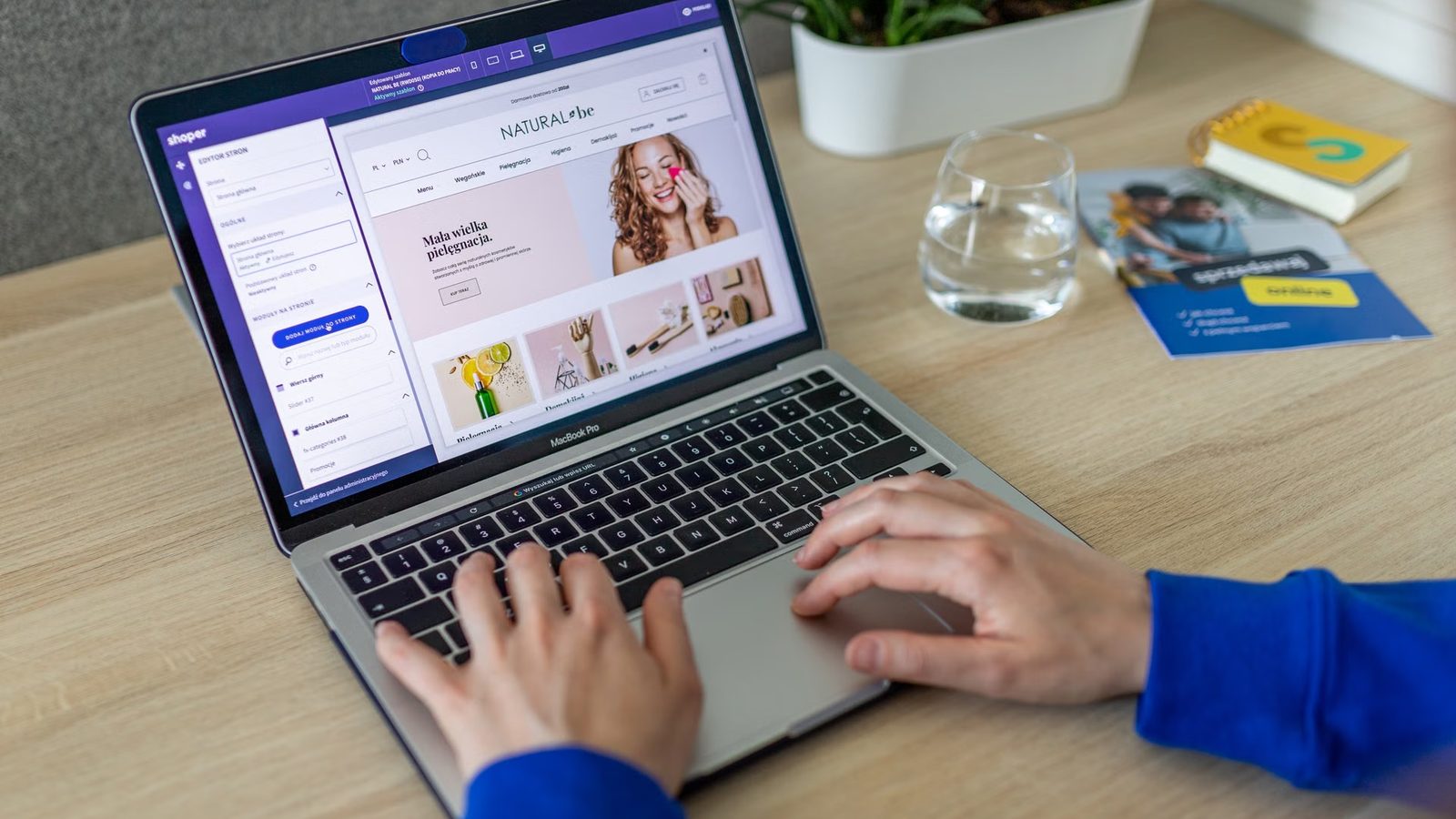 Dłonie na klawiaturze srebrnego laptopa, na ekranie panel administracyjny sklepu internetowego i podgląd strony sklepu z kosmetykami, obok szklanka wody i doniczka z rośliną