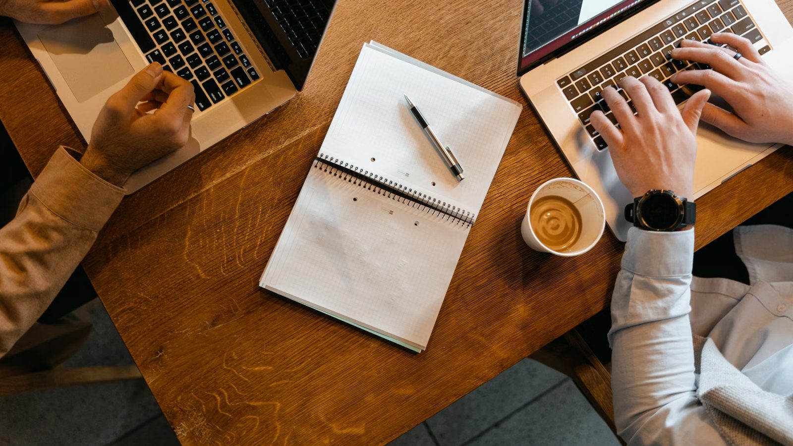 Dwie osoby przy laptopach nad wspólnym stołem, pośrodku otwarty notes z długopisem
