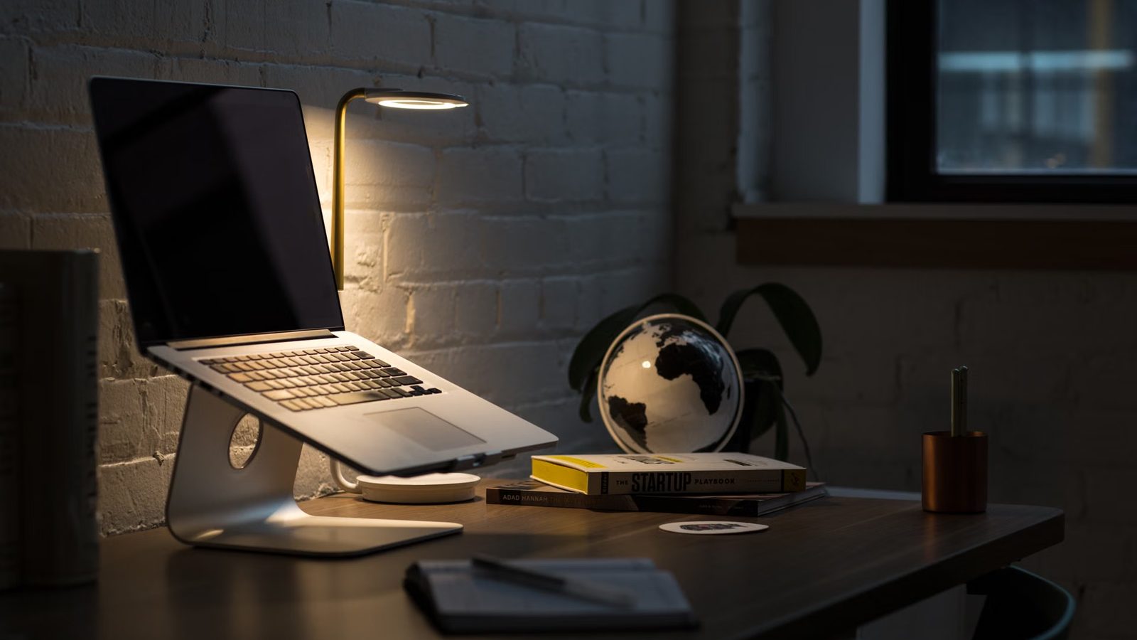 Laptop na podstawce i zapalona lampka na drewnianym biurku, obok globus, stos książek i notes