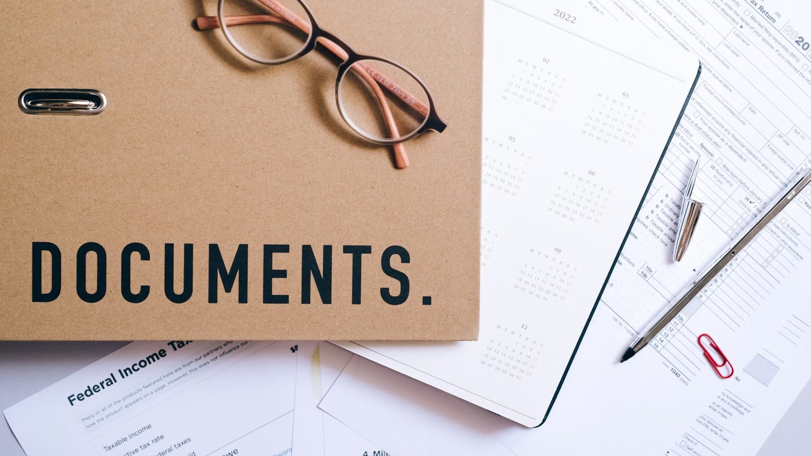 Okulary na brązowej teczce z napisem DOCUMENTS, obok kalendarz, notes, długopis i wydrukowane formularze