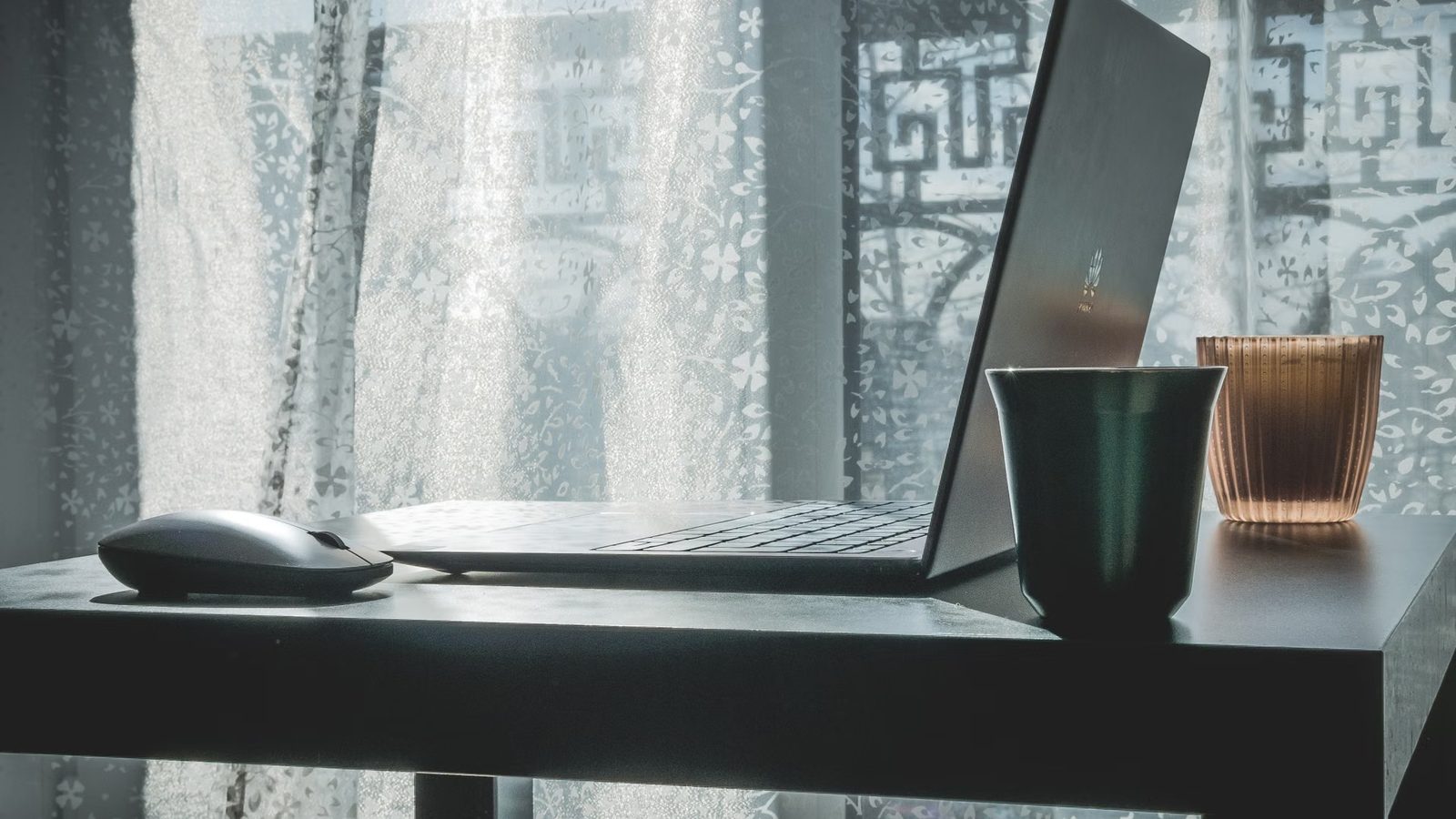 Otwarty laptop, mysz i dwa kubki na ciemnym biurku przy oknie z firanką
