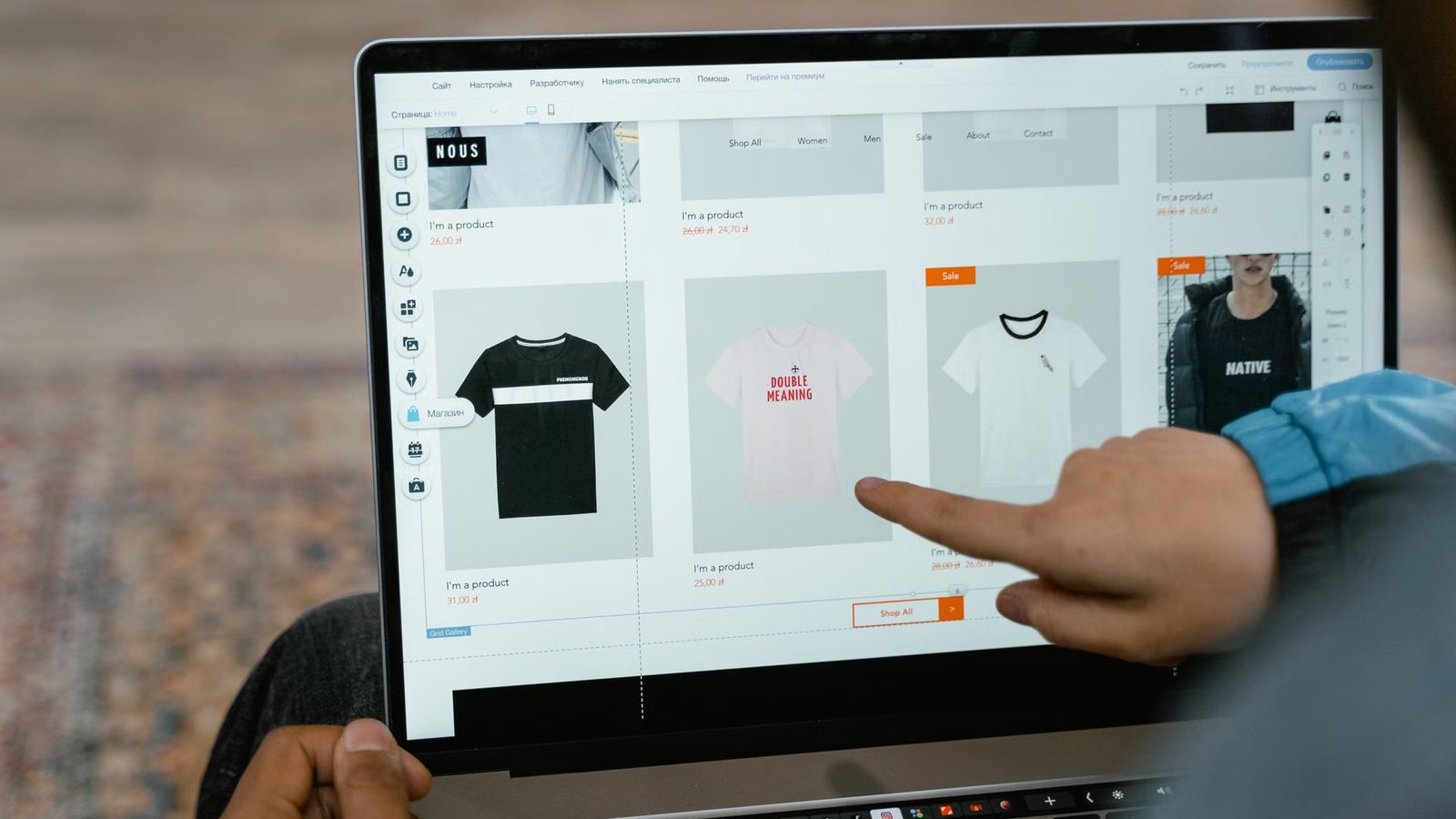 Palec wskazuje kafelek z koszulką w siatce kart produktów otwartej w edytorze sklepu na ekranie laptopa