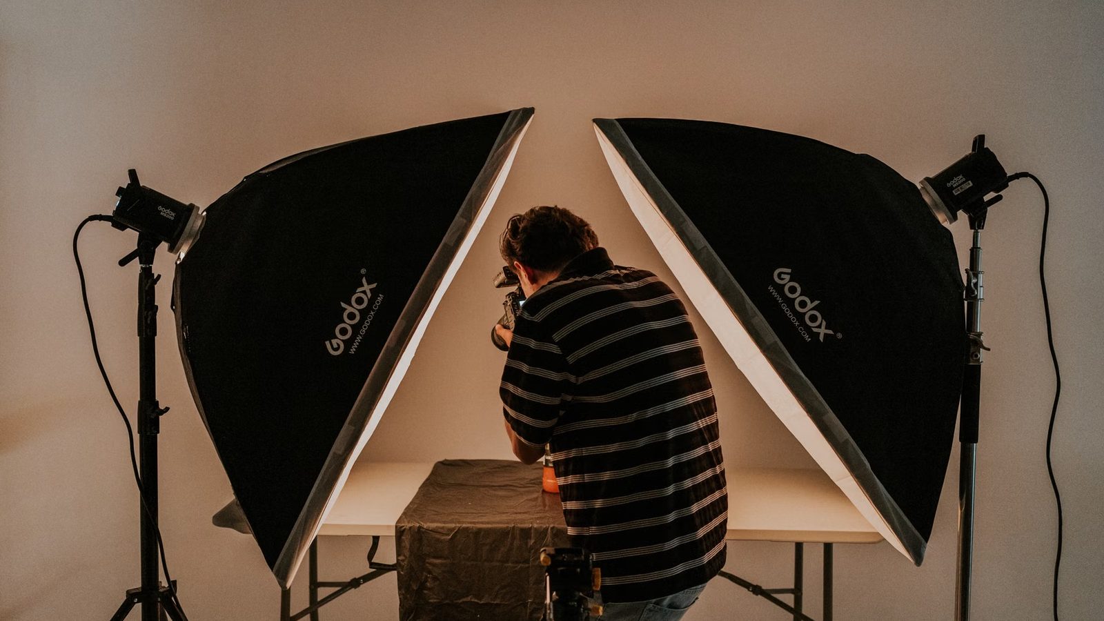 Fotograf pochylony nad białym stolikiem między dwiema lampami studyjnymi z softboksami