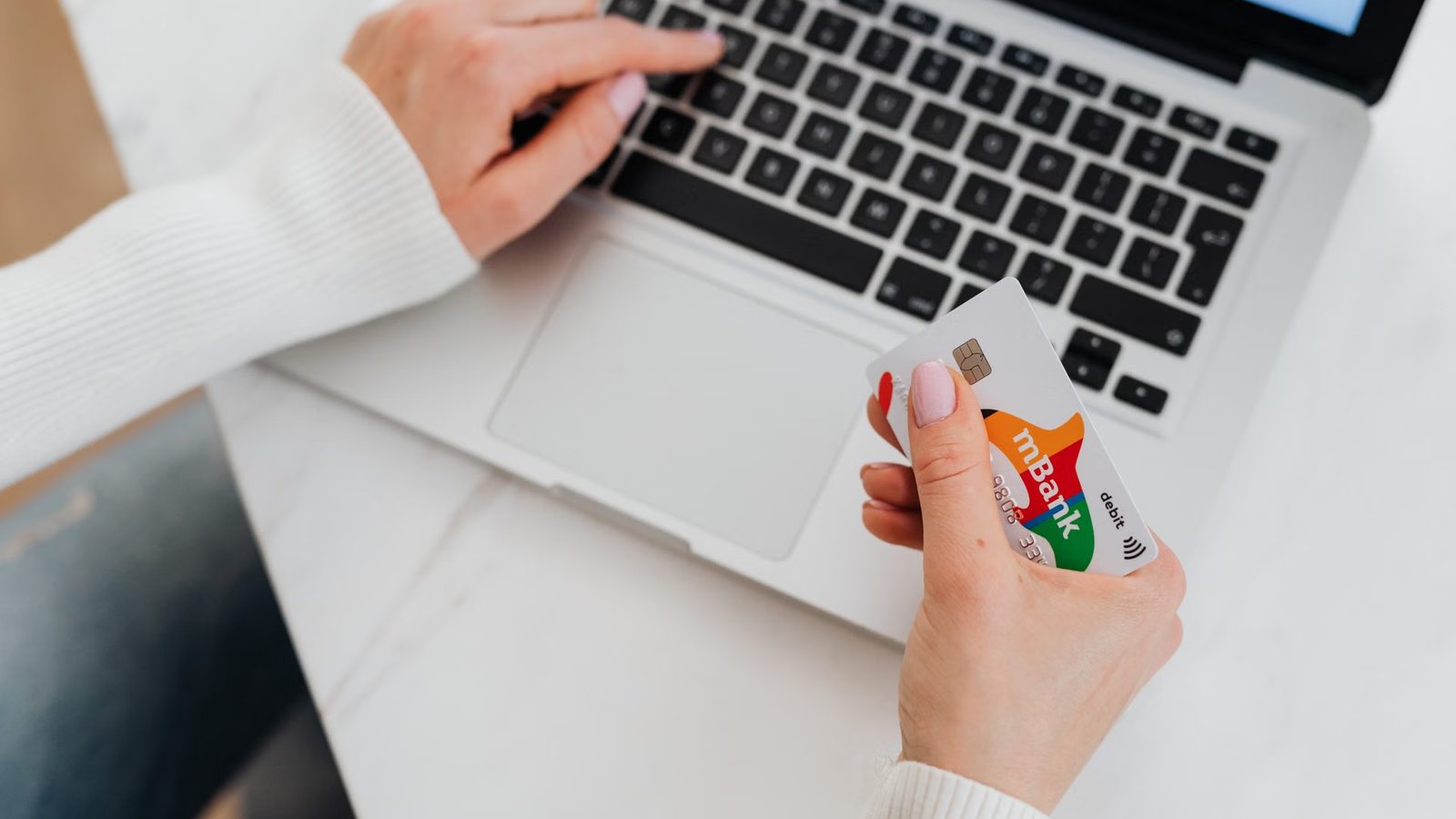Dłoń na klawiaturze laptopa, w drugiej ręce karta płatnicza