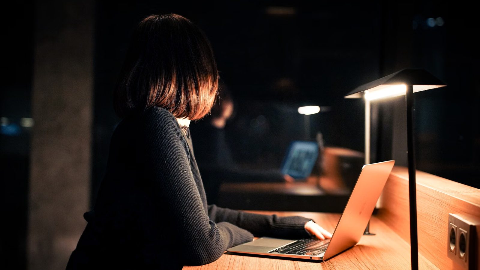 Osoba przy laptopie w ciemnym pomieszczeniu, oświetlona lampką biurkową stojącą na drewnianym blacie