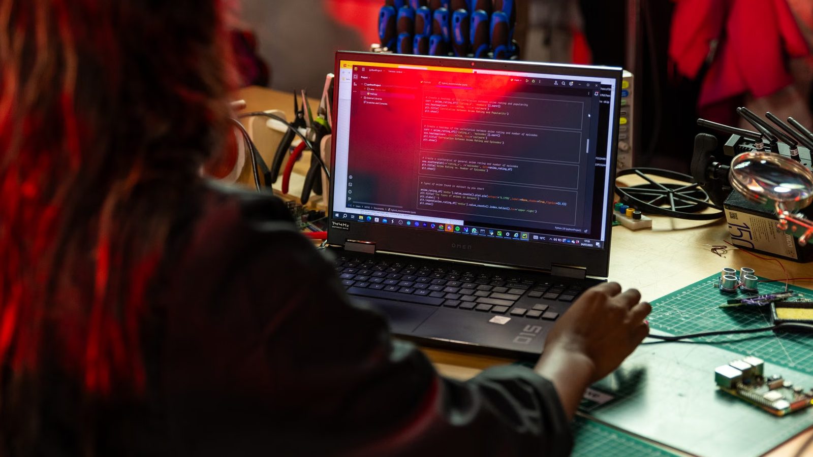 Osoba przy laptopie z otwartym edytorem kodu, obok lupa i elektronika na stole warsztatowym