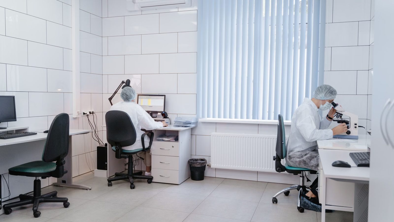 Dwie osoby w czepkach i fartuchach pracują w laboratorium, jedna przy mikroskopie, druga przy komputerze