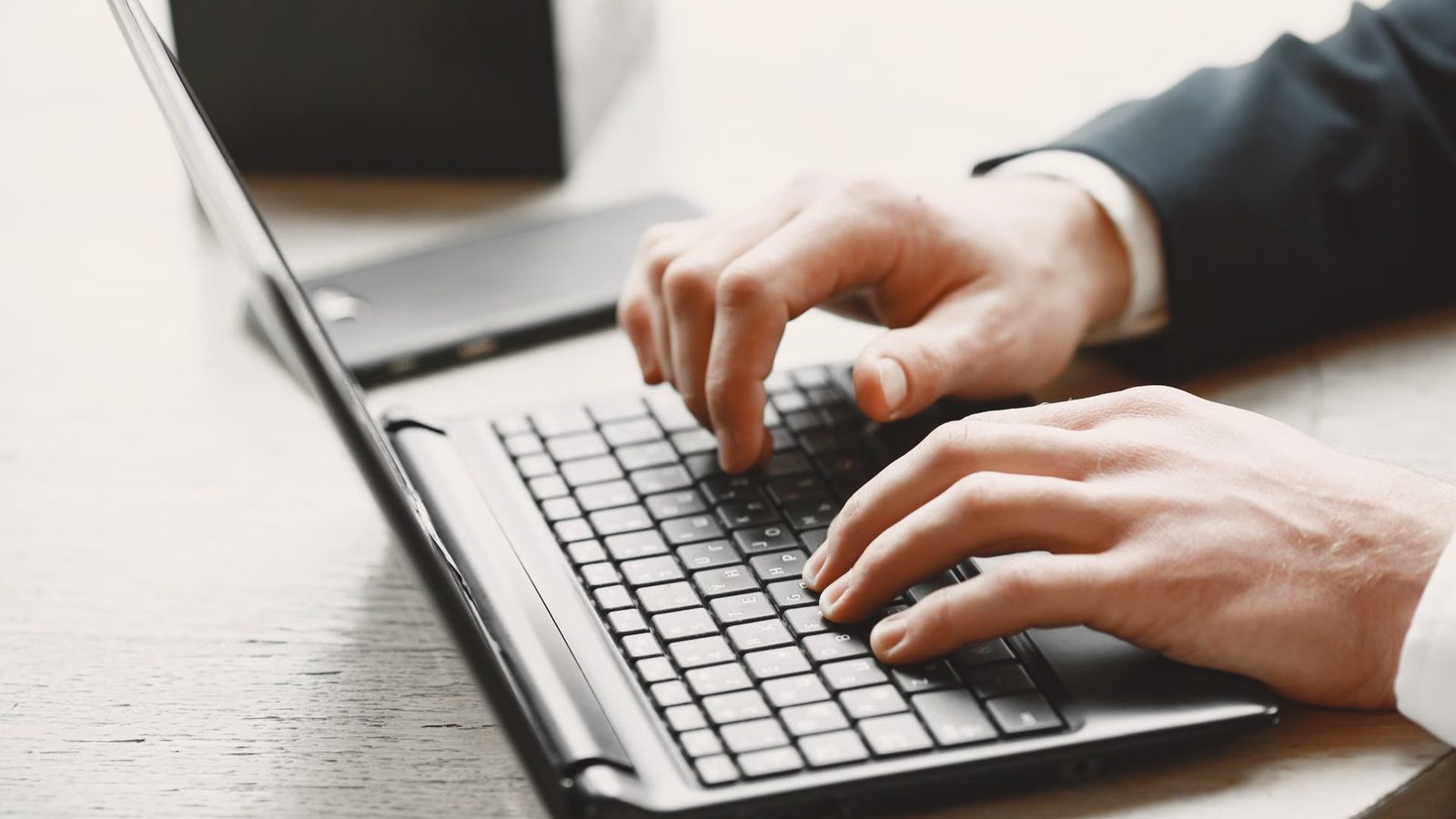 Dłonie mężczyzny w koszuli piszą na klawiaturze laptopa na jasnym biurku