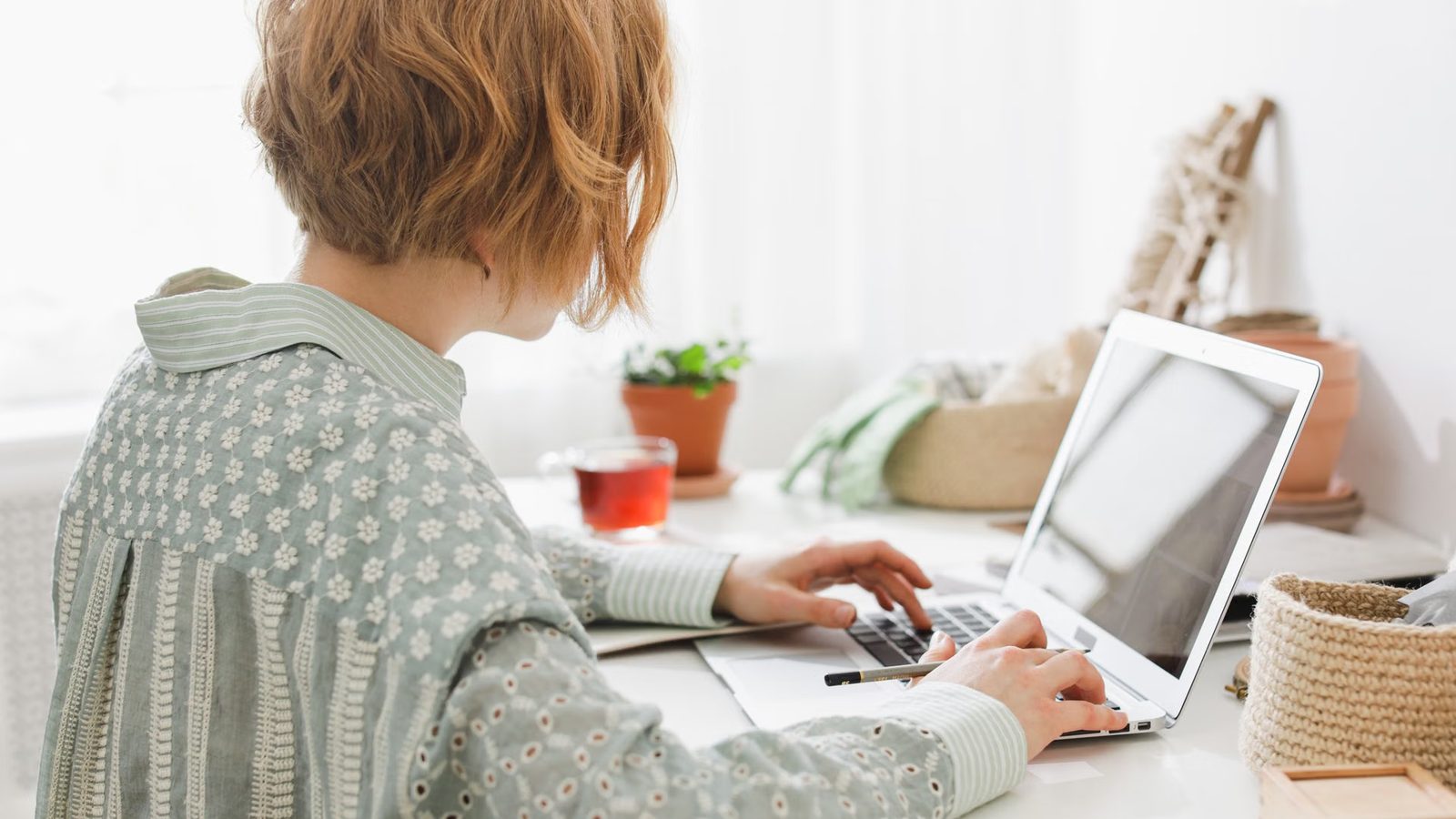 Kobieta pisze na laptopie przy białym biurku w jasnym pokoju, obok szklanka herbaty