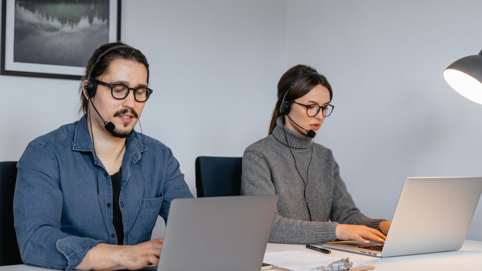 Mężczyzna i kobieta w zestawach słuchawkowych pracują przy laptopach na białym biurku z lampą