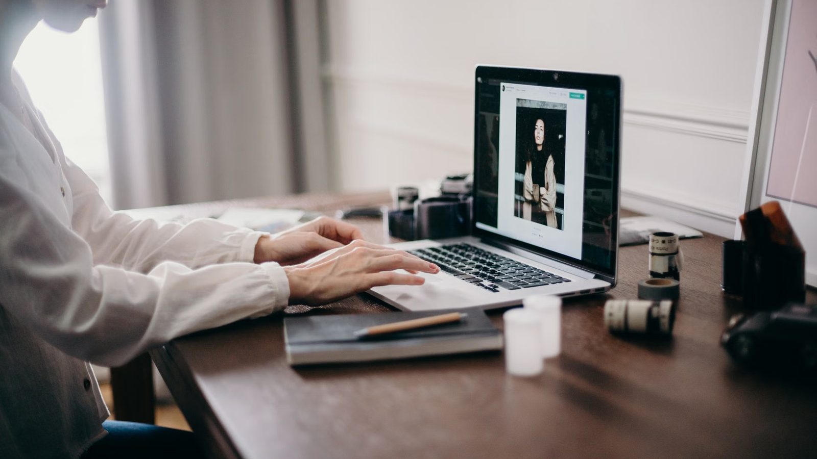 Dłonie na klawiaturze laptopa, obok notes, ołówek i rolki filmu na drewnianym biurku