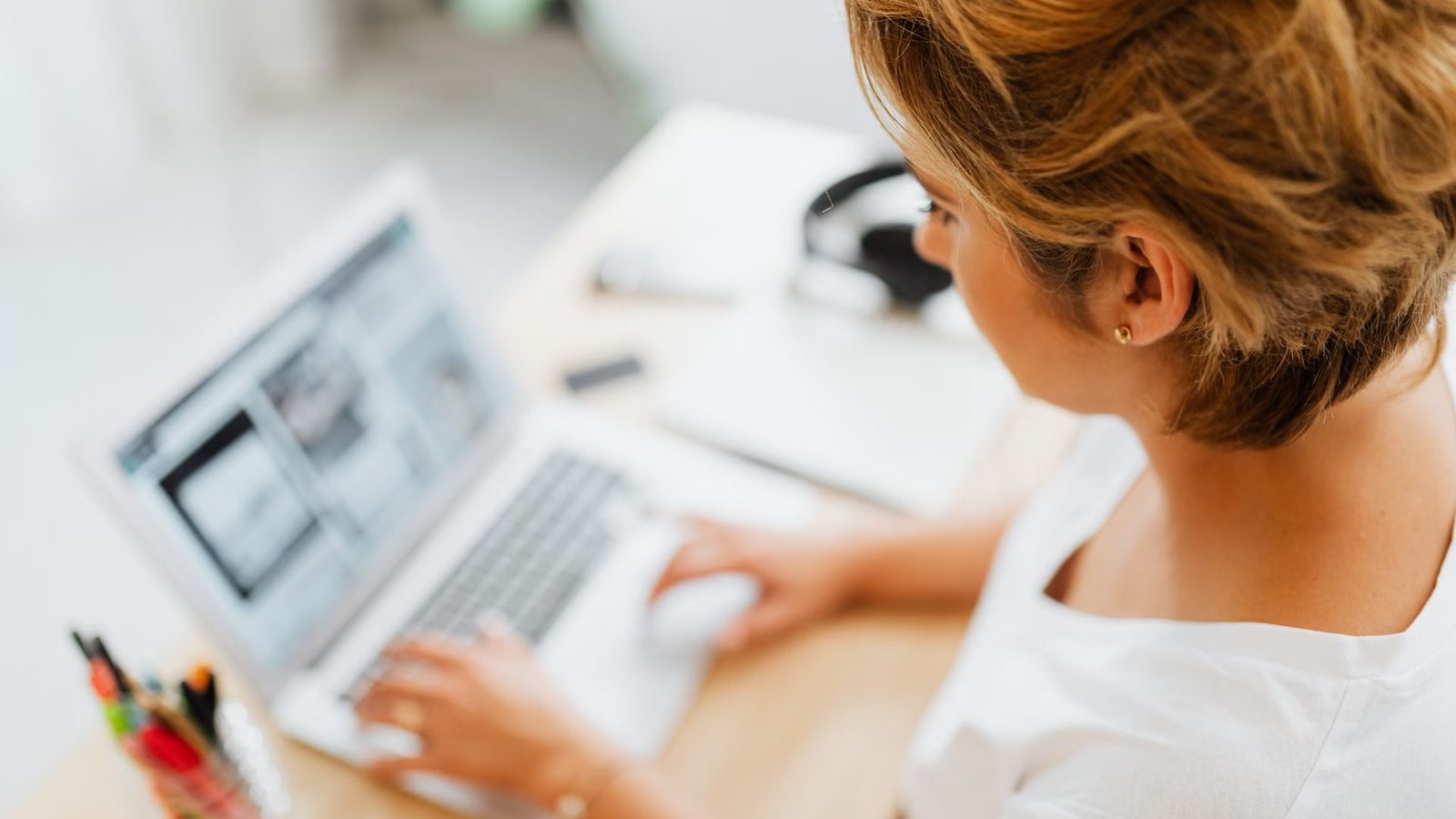 Kobieta pracuje na laptopie przy jasnym biurku, obok słuchawki i kubek z długopisami