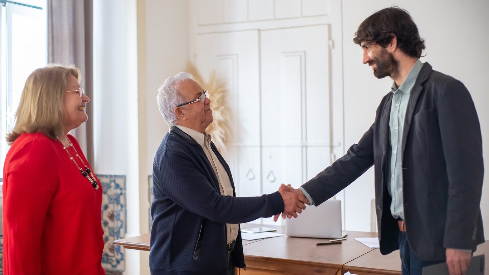 Dwóch mężczyzn podaje sobie dłonie przy biurku z laptopem, obok stoi kobieta w czerwonym ubraniu