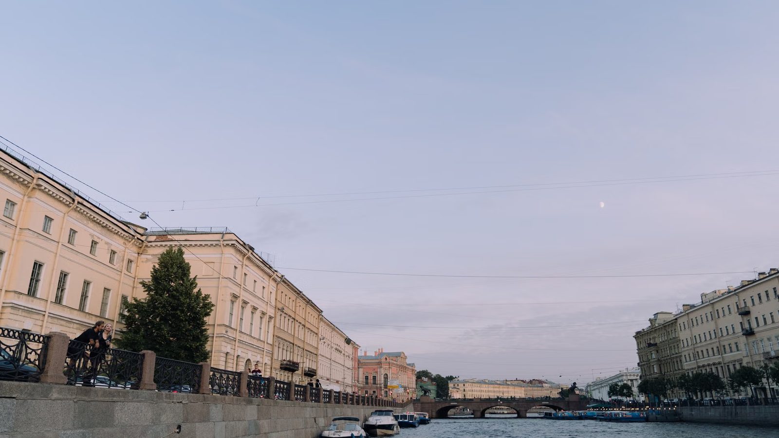 Kamienice wzdłuż nabrzeża rzeki z łodziami przy brzegu i kamiennym mostem w oddali