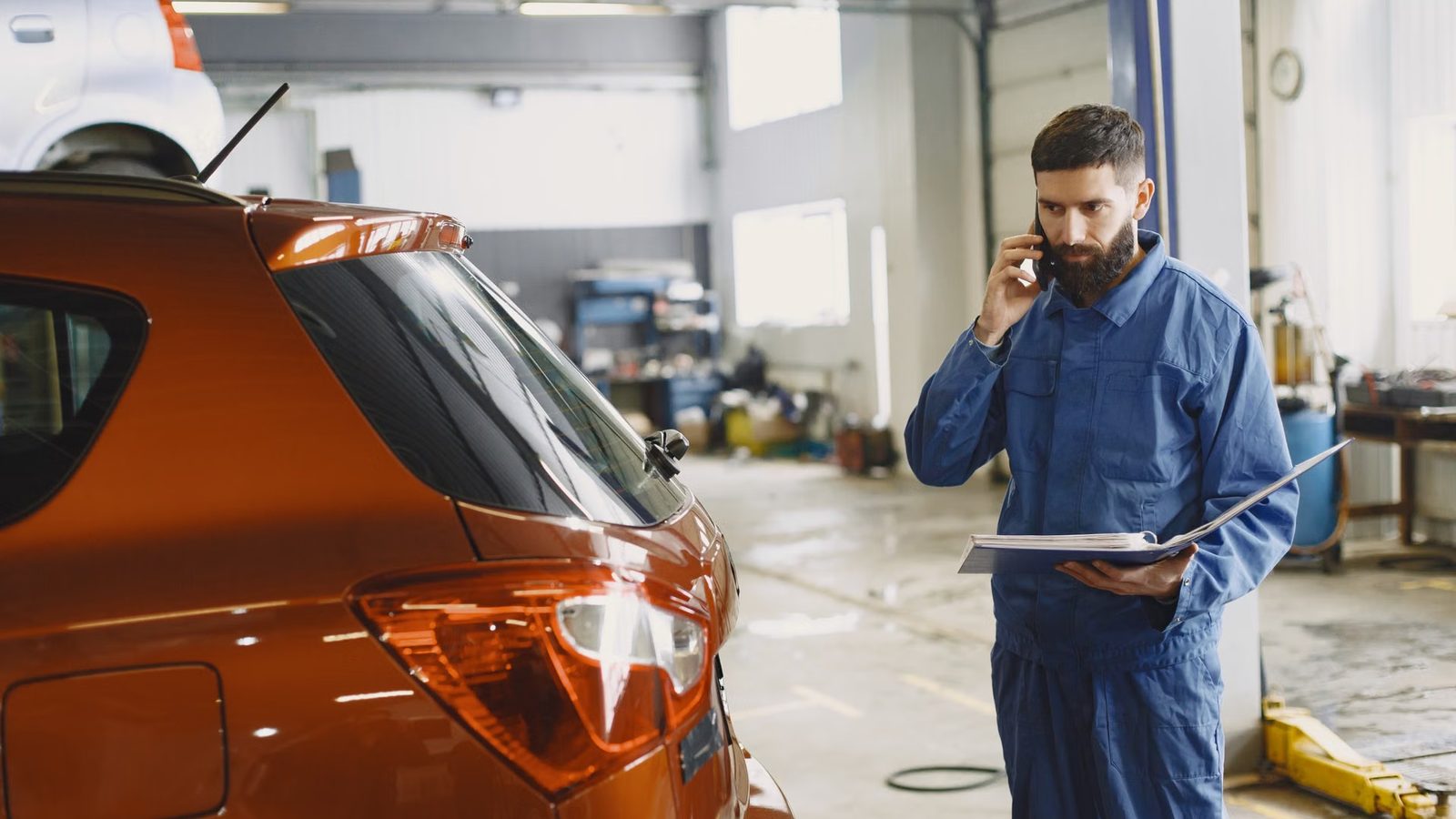 Mechanik w kombinezonie rozmawia przez telefon obok auta w warsztacie
