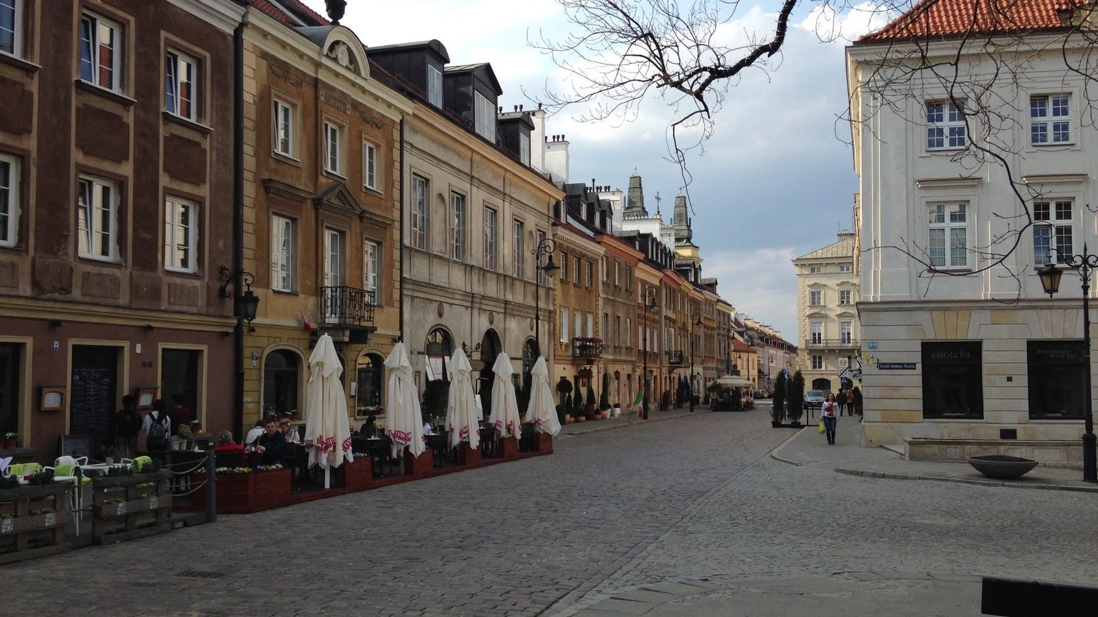 Pustawa brukowana ulica starego miasta z rzędem zabytkowych kamienic, złożonymi parasolami ogródka i pojedynczymi przechodniami w głębi