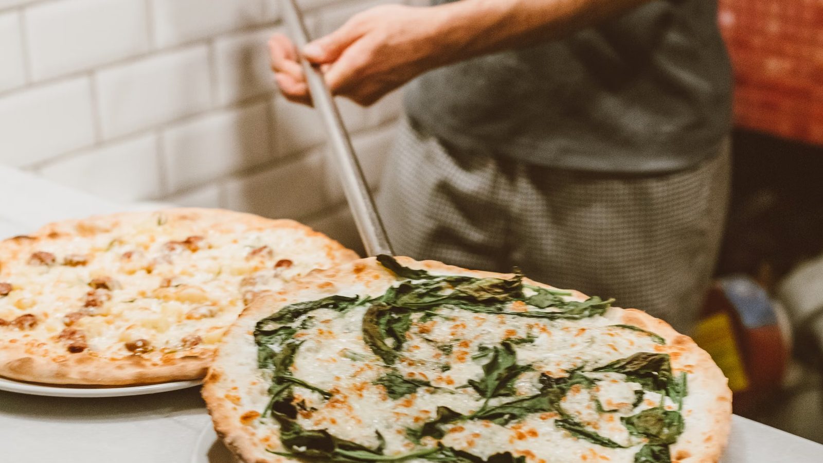 Dwie pizze leżące na blacie, za nimi kucharz z łopatą do pizzy