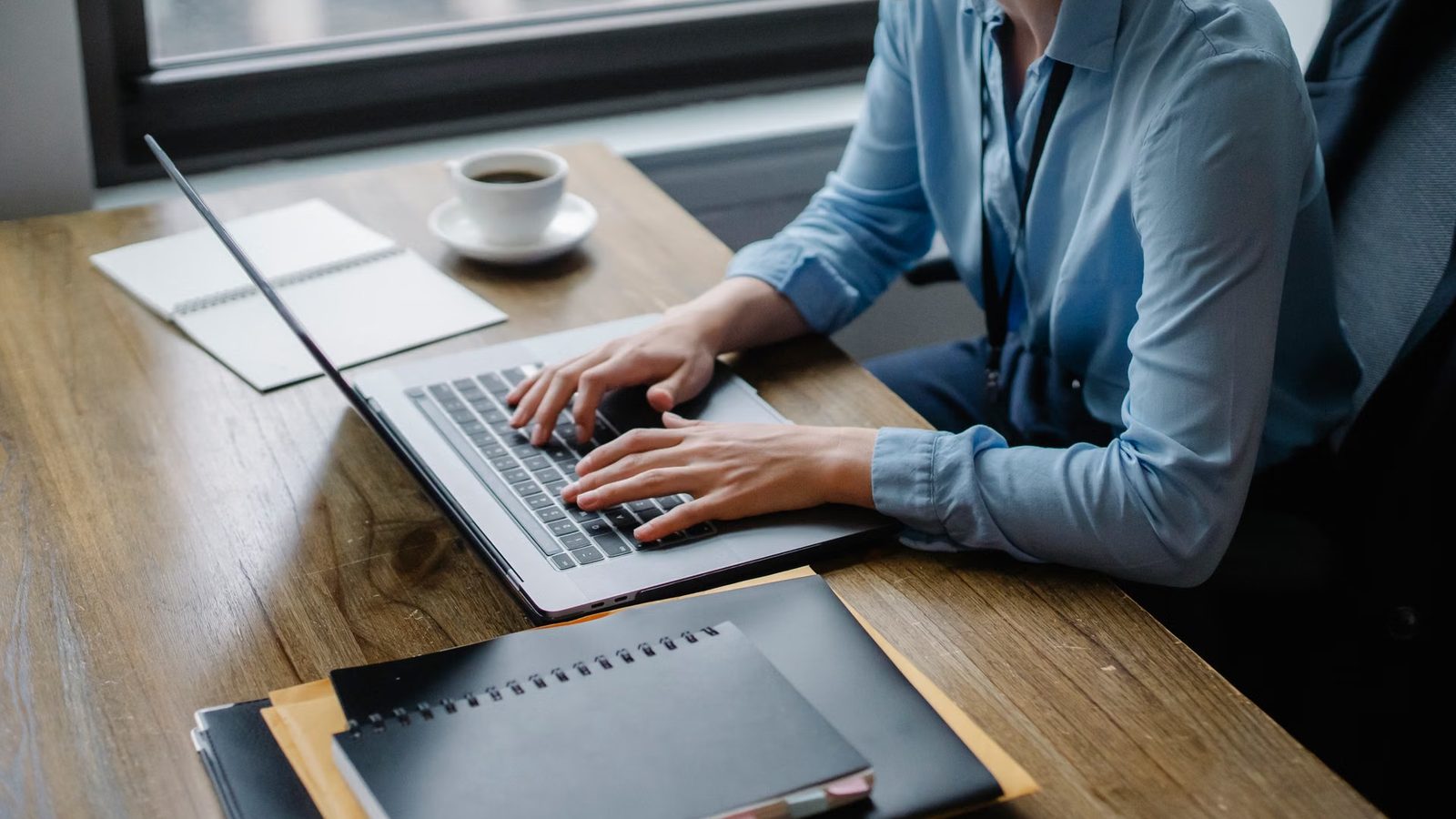 Osoba w niebieskiej koszuli pisze na laptopie przy drewnianym biurku z notesami i kawą