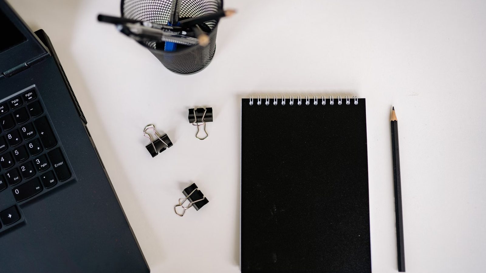 Laptop, czarny notes, ołówek i spinacze biurowe na białym biurku